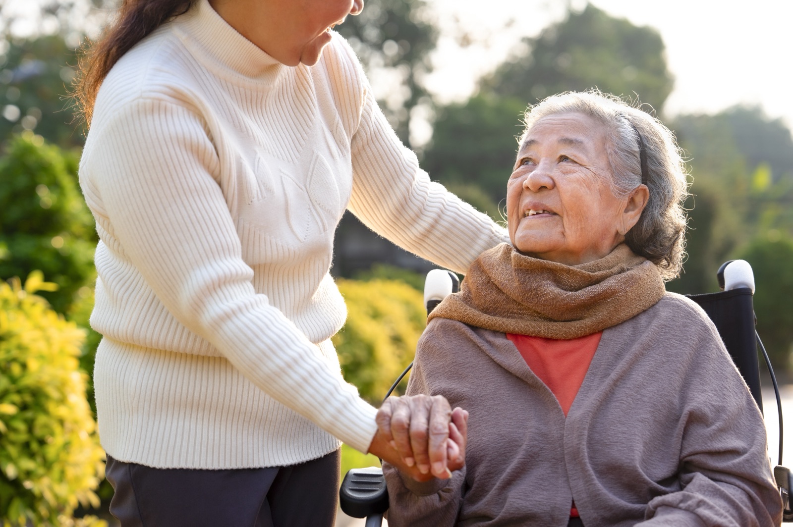 An older woman in a wheelchair holding another woman’s hand in a garden.