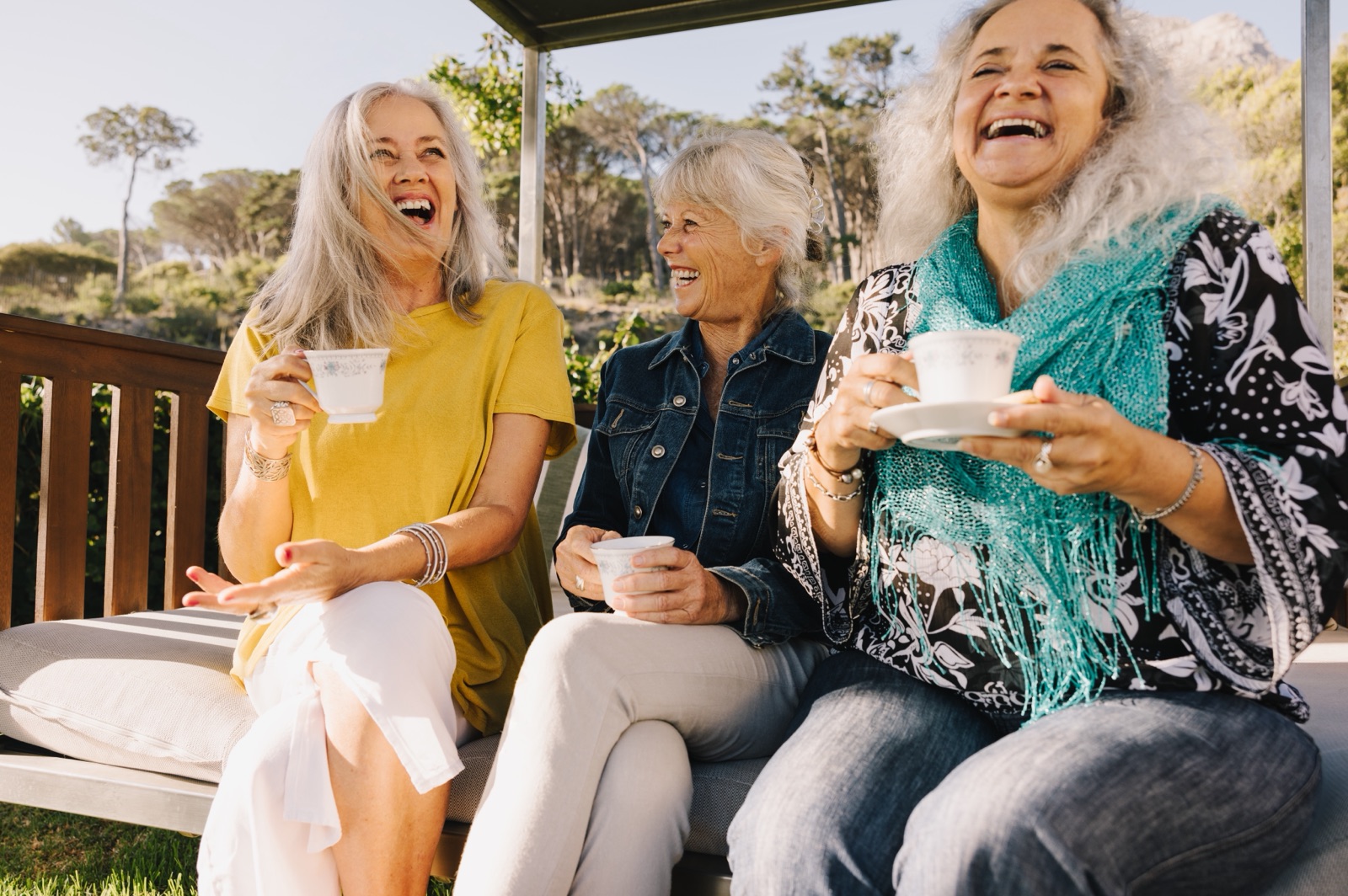 Three older women laughing together with cups of tea outdoors.