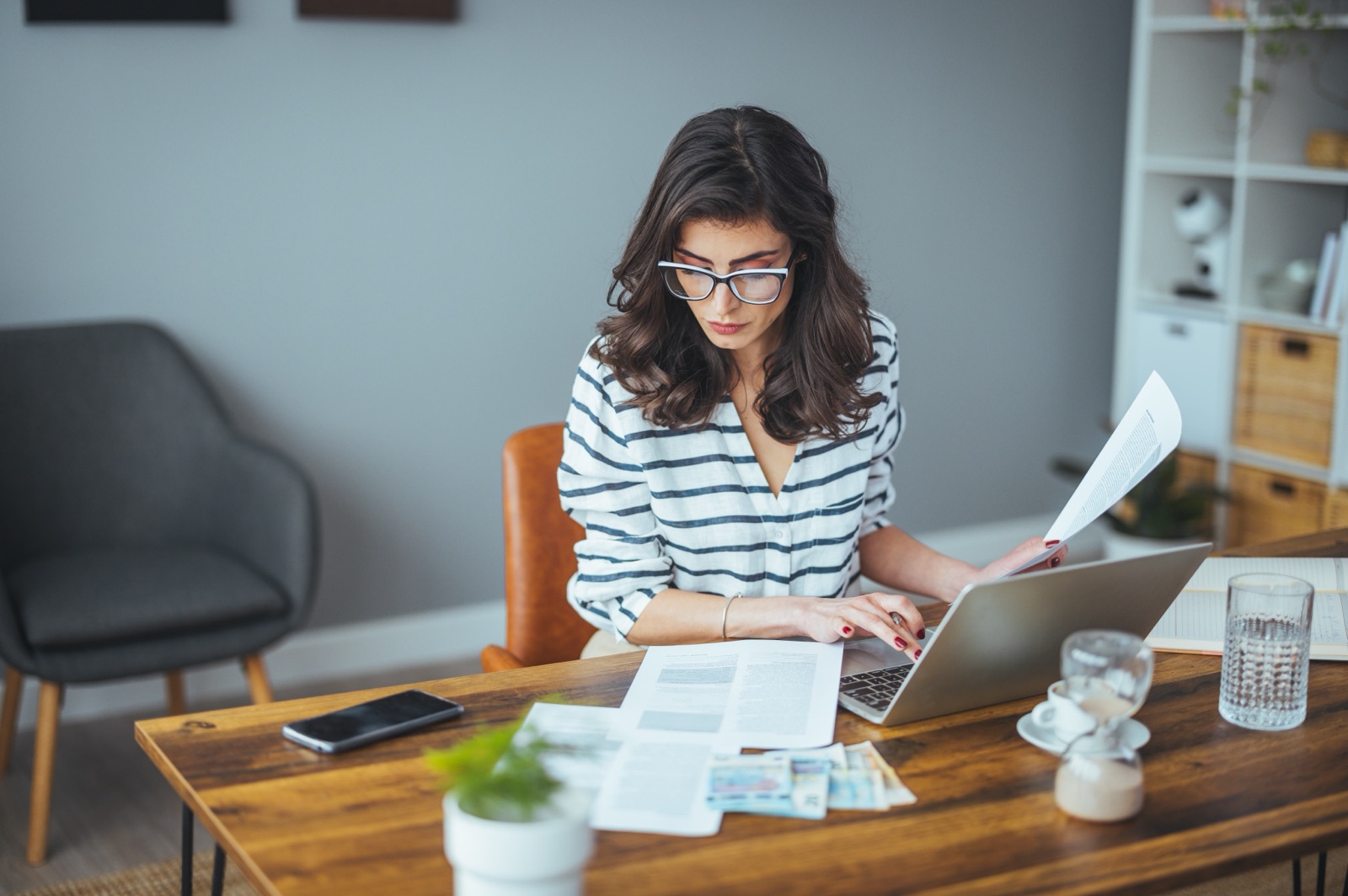 A woman reviewing paperwork while using a laptop at a table.