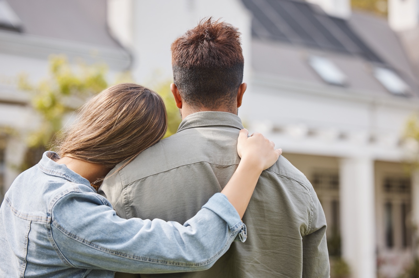 A couple embracing while looking at a house, viewed from behind.
