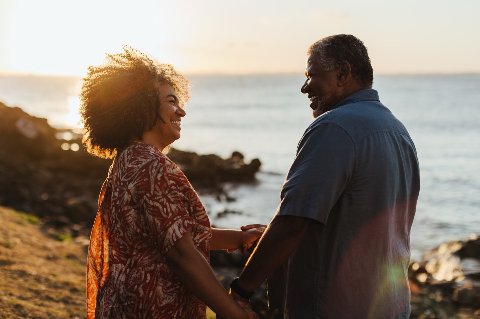 An older couple holding hands beside the sea at sunset.